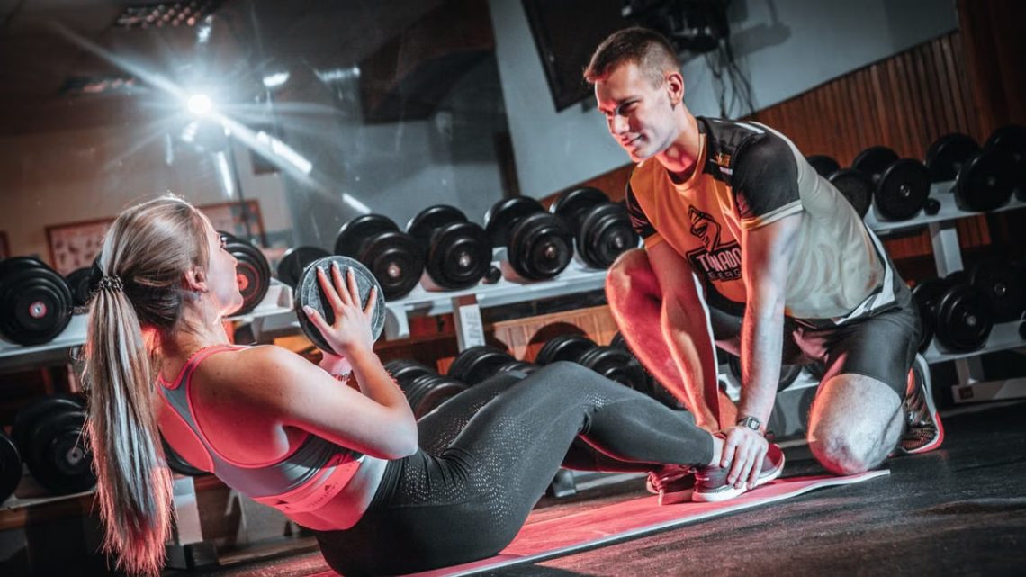 A personal trainer assists a woman doing sit-ups with a medicine ball in a gym setting.