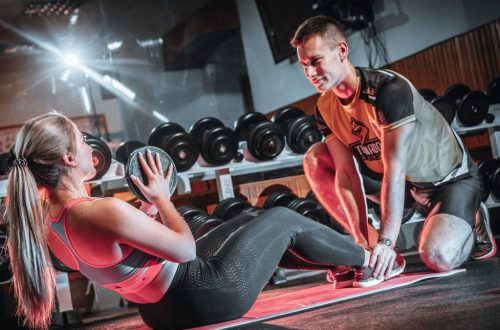 A personal trainer assists a woman doing sit-ups with a medicine ball in a gym setting.