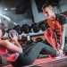 A personal trainer assists a woman doing sit-ups with a medicine ball in a gym setting.
